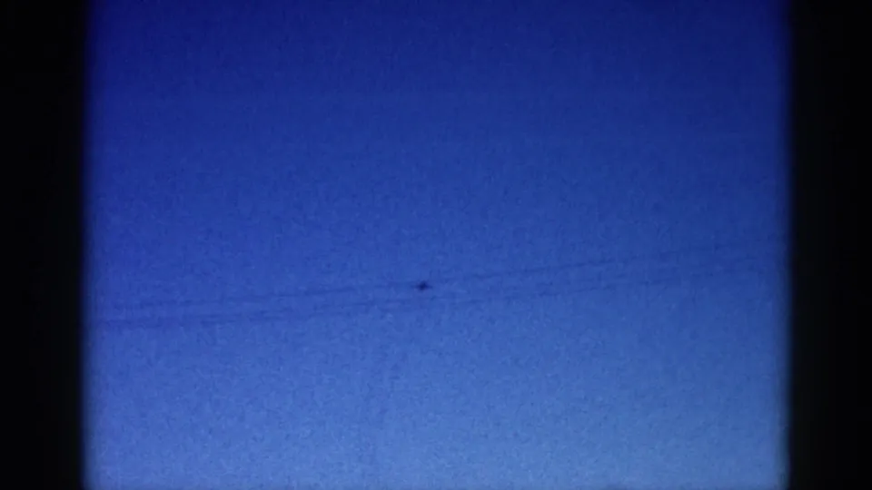8mm archival frame circa 1962 showing deep blue dusk sky with a distant aircraft silhouette, faint possible power line, and period film grain.