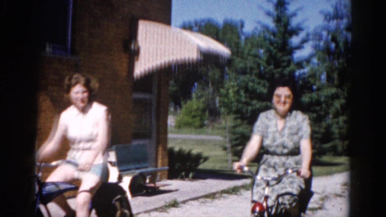 Two women cycling on a suburban driveway in what appears to be early 1960s Illinois, with a brick house facade and striped canvas awning visible behind them.