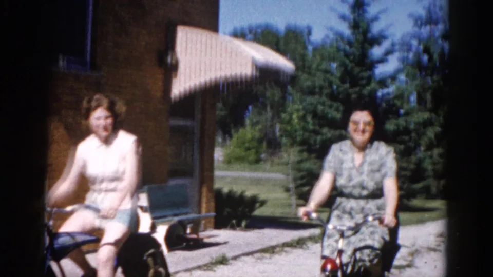 Two women cycling on a suburban driveway in what appears to be early 1960s Illinois, with a brick house facade and striped canvas awning visible behind them.
