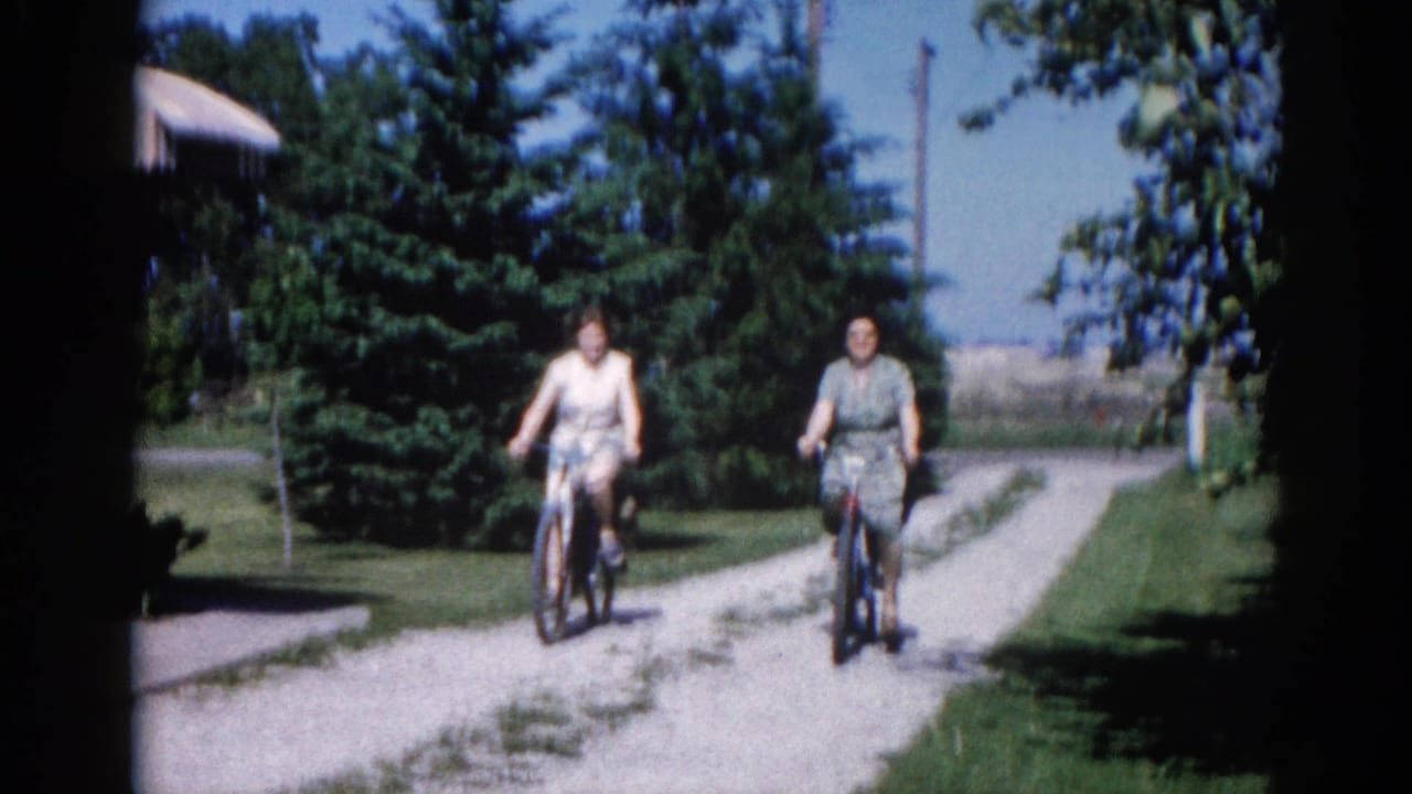 Two women riding bicycles toward camera on a residential gravel path; tall conifers and clear sky behind them.