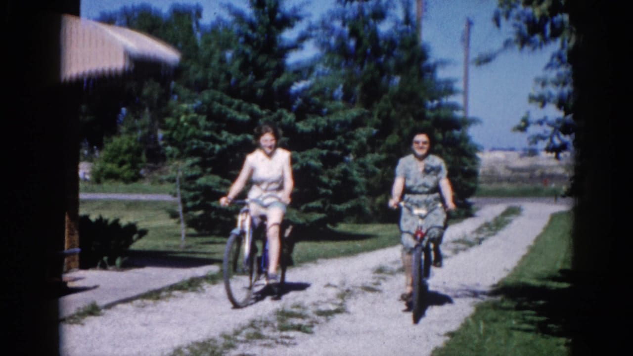 Two women cycling toward camera on a residential gravel path, circa 1962 Illinois; broader sky and faint open landscape visible behind the conifer row.