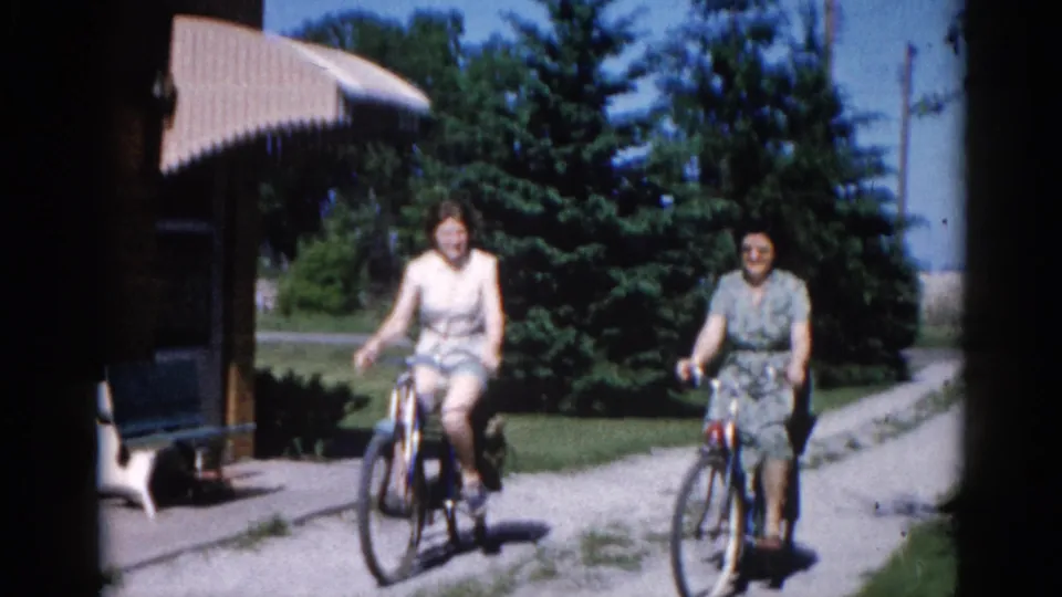 Two women on bicycles ride close toward camera on a residential gravel path, circa 1962 Illinois; a small white animal is visible at the lower-left frame edge.