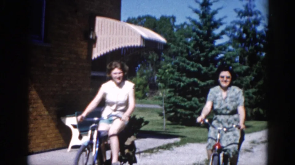 Two women cycling directly toward camera in close view, circa 1962 Illinois; brick building at right frame edge, one woman in sunglasses and a floral teal dress.