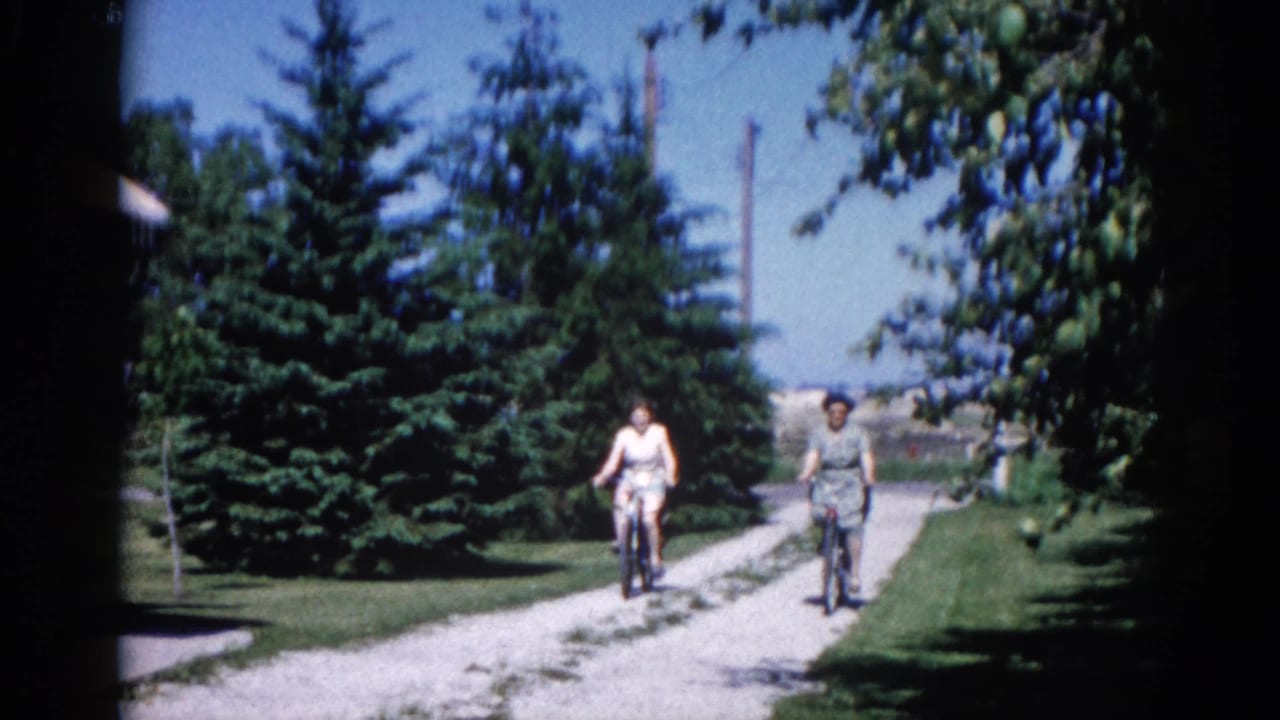 Two women cycling side-by-side on a conifer-lined gravel lane, faces becoming visible; 8mm home movie, Illinois circa 1962.