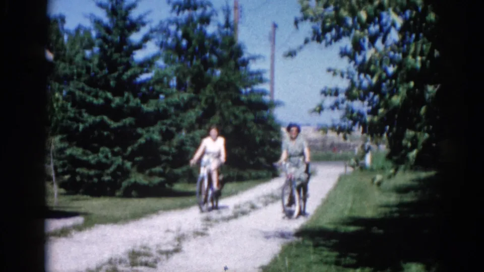 Two women in 1960s day dresses cycling on a gravel lane, faces clearly visible; 8mm home movie, Illinois circa 1962.