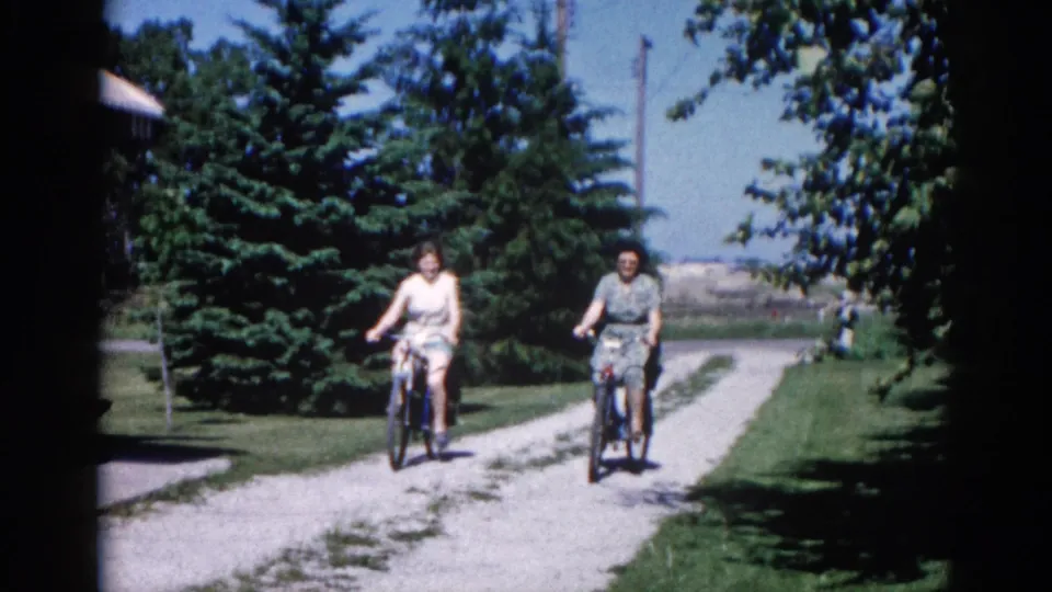 Close view of two women cycling on a conifer-lined gravel lane in day dresses; 8mm home movie, Illinois circa 1962.