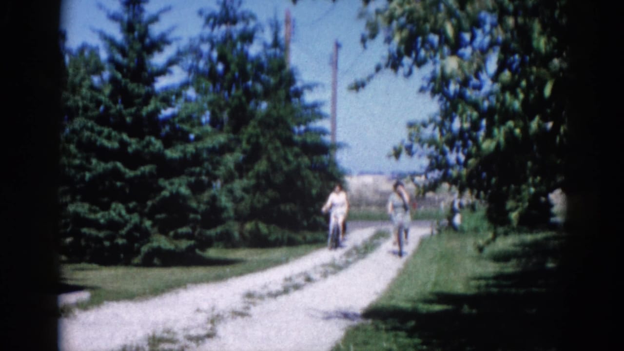 Two cyclists on bicycles approach along a gravel path lined with coniferous trees, slightly closer than the opening frame.