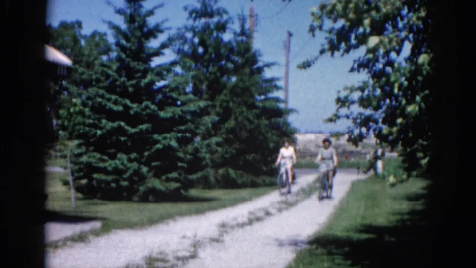 Two cyclists in casual dress approaching camera on a gravel path with a partially visible building at left and deciduous branches overhead; Illinois, circa 1962 per metadata.