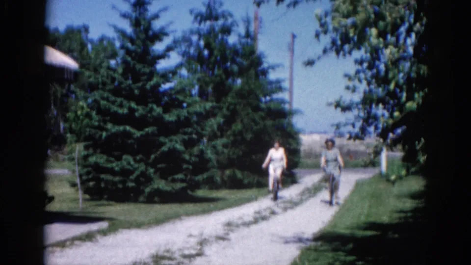 Two cyclists passing the fixed camera position at close range on a sunlit gravel path; evergreen trees and a partially visible building frame the scene; Illinois, 1962 per metadata.