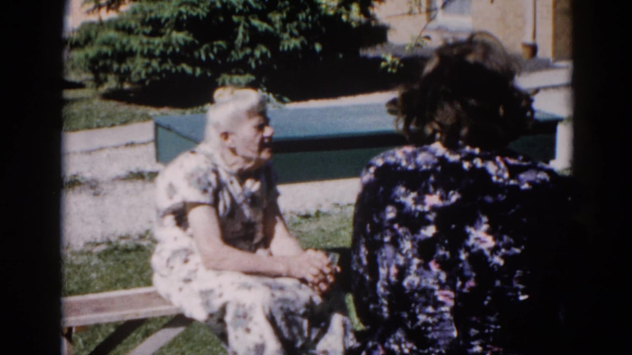Two women in patterned dresses seated on a wooden bench, conversing outdoors on a sunny day.