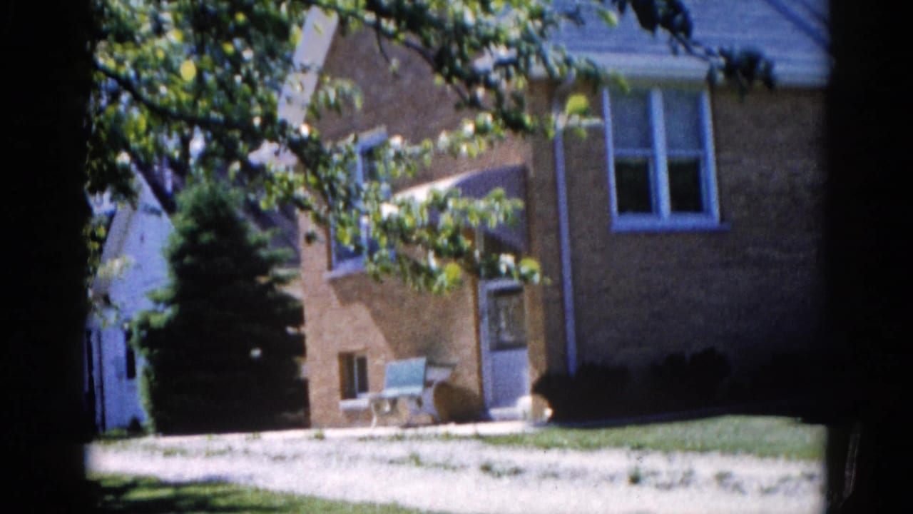Exterior of a two-story tan brick house with white-trimmed windows, green lawn, and surrounding mature trees.