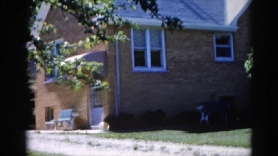 Tree-framed view of a tan brick house with white-trimmed windows and outdoor chairs on a sunny front lawn, Illinois, circa 1962.