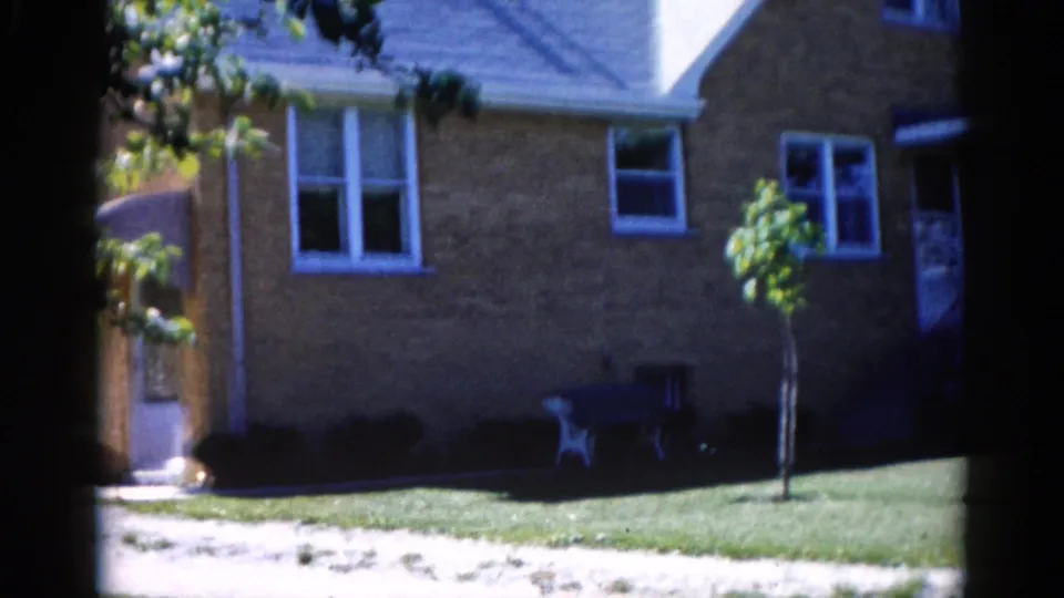 Tan brick suburban house with white-trimmed windows, a young sapling, and outdoor chairs on the front lawn, Illinois, circa 1962.