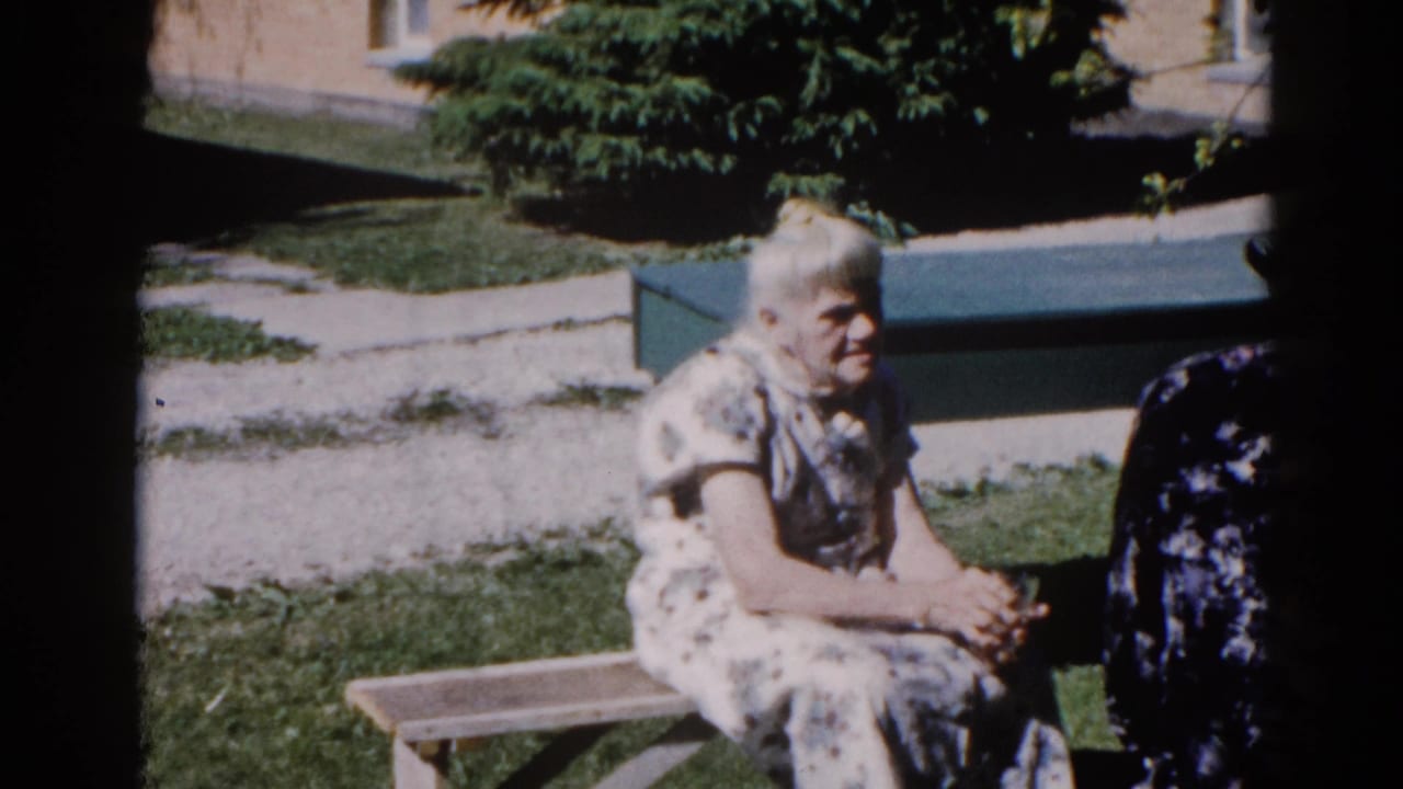Elderly white-haired woman in a white floral dress sits on a wooden bench outdoors; a second person in a dark floral dress is partially visible beside her.