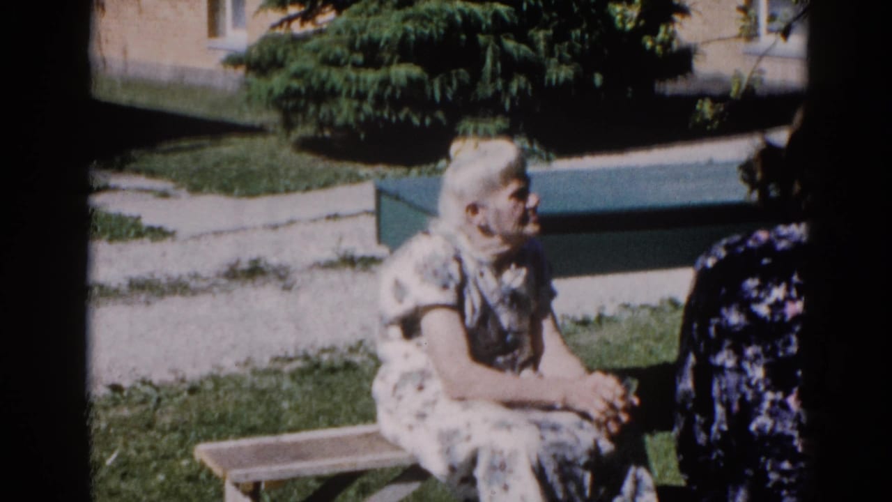 Elderly woman in white floral dress mid-conversation on a garden bench, Illinois, circa 1962; second figure in dark floral dress visible at right edge.