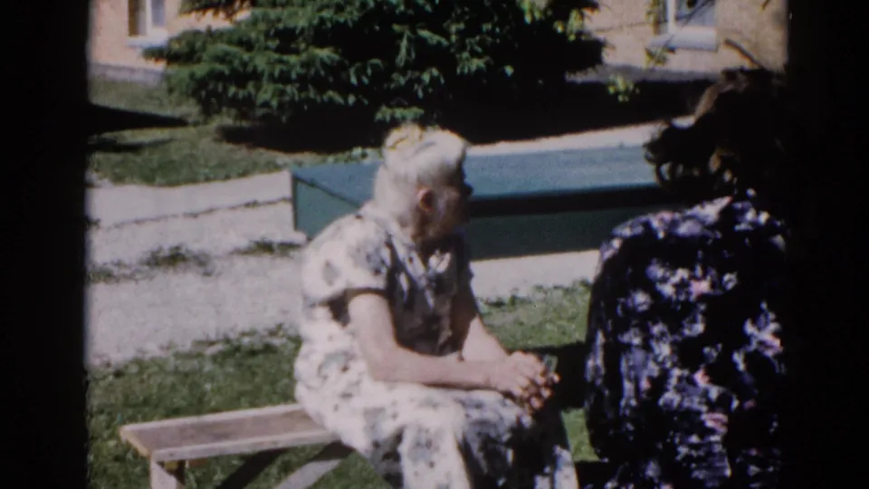 Over-shoulder view of two women in outdoor conversation, Illinois, circa 1962; the elderly woman on the bench looks toward the dark-dressed figure in the foreground.