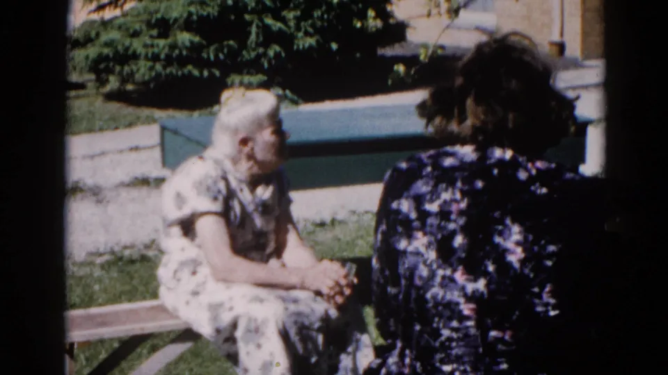 Elderly woman in white floral dress leans in conversation toward a companion, garden bench, Illinois, circa 1962; warm 8mm color tones visible.