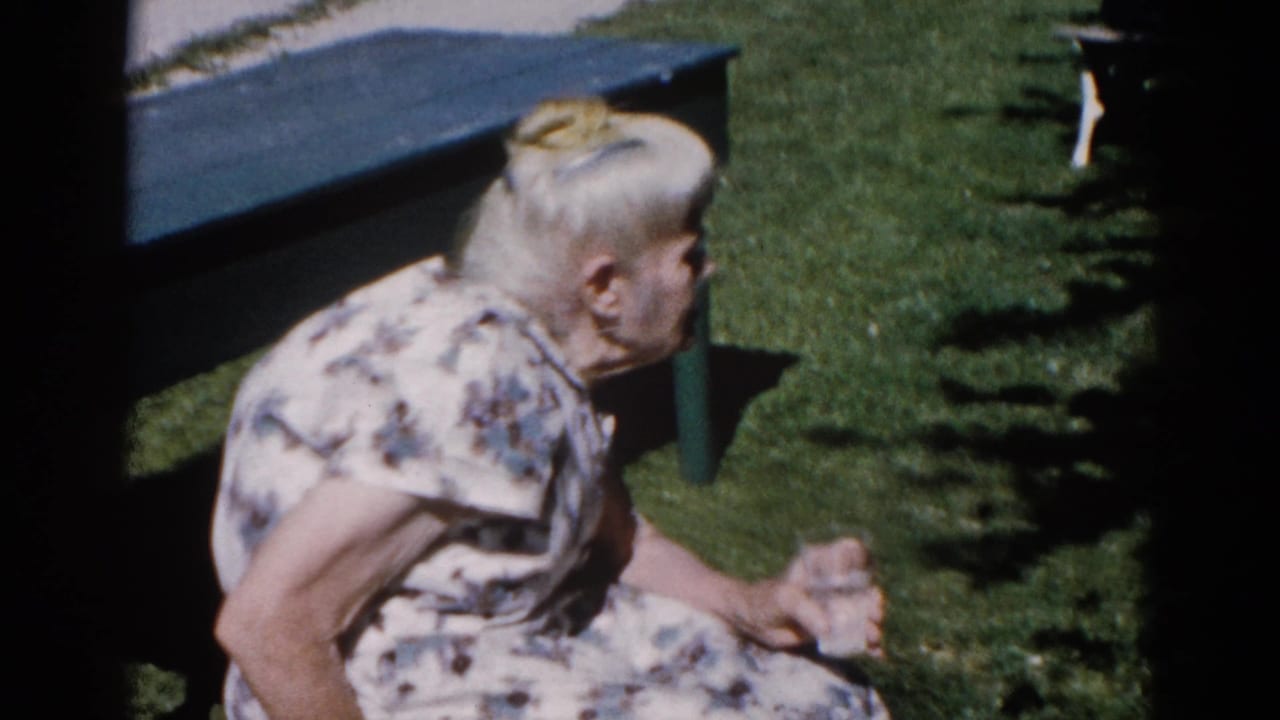 Older woman in white floral dress seated in profile on a painted garden bench, 8mm home movie, Illinois circa 1962.