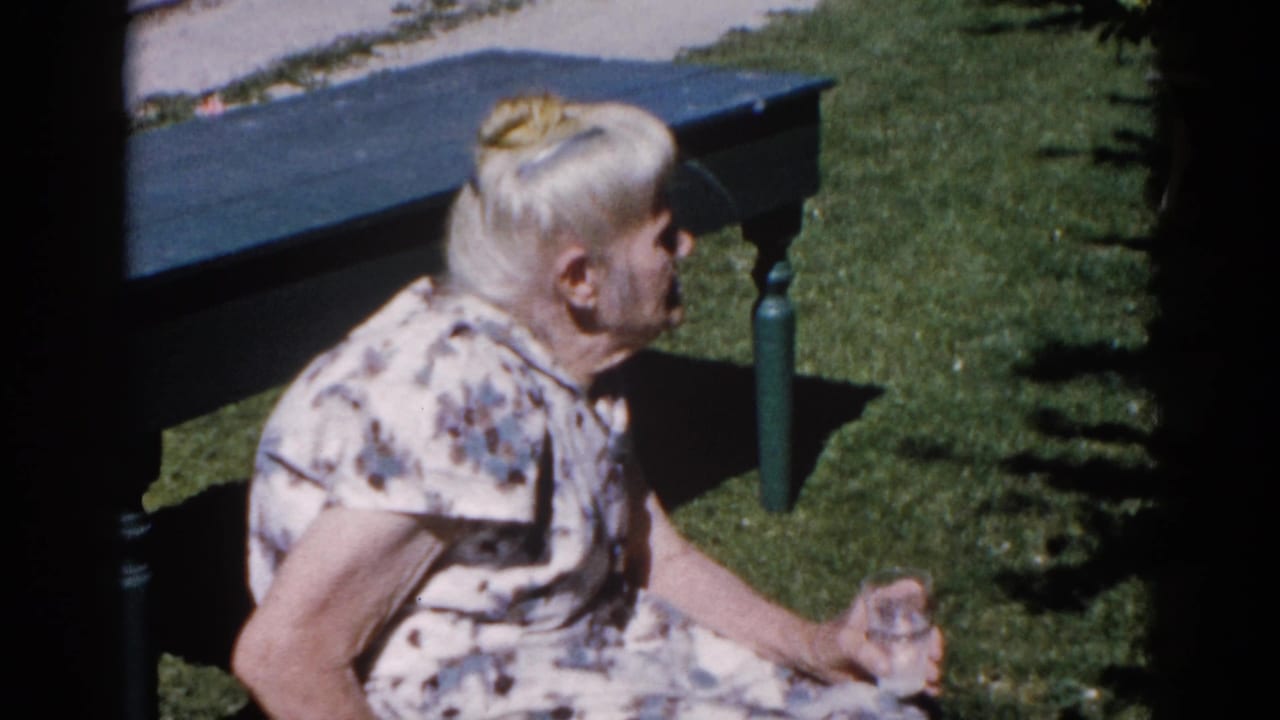 Older woman in floral house dress holding a glass on a painted garden bench, small evergreen shrub in background, 8mm home movie Illinois circa 1962.