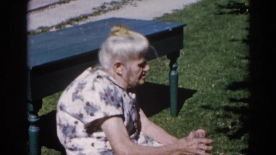 Older woman in white floral house dress turning toward camera on a painted garden bench, sunlit lawn background, 8mm home movie Illinois circa 1962.