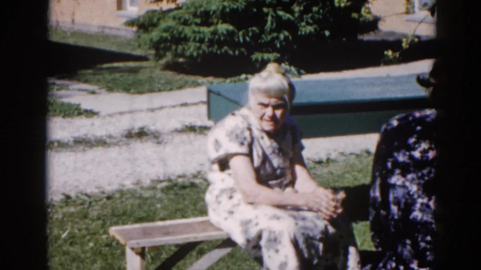 Two women outdoors in a garden setting — one older in white floral dress on a bench, a second figure partially in frame at right — 8mm home movie Illinois circa 1962.