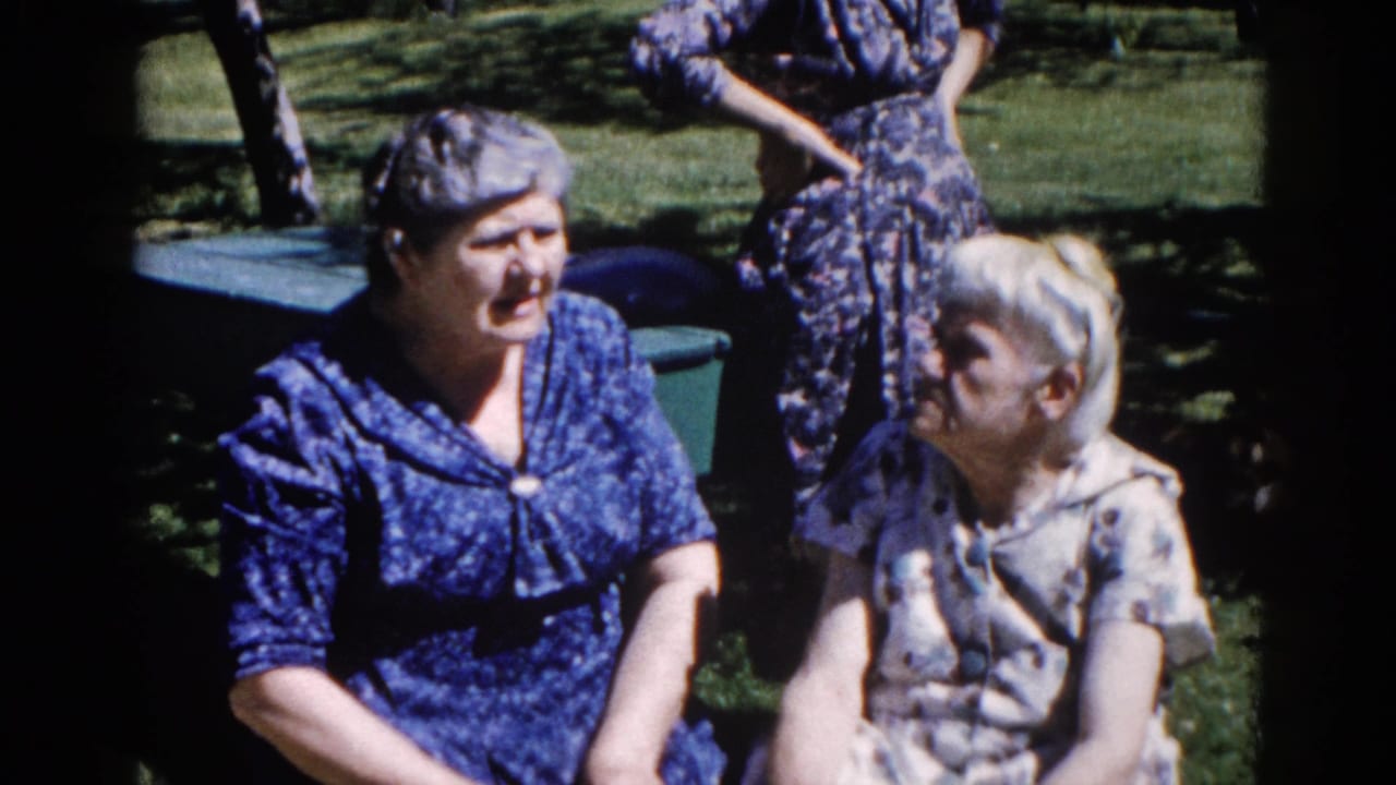 Two older women in early-1960s patterned dresses seated on a sunlit lawn in Illinois, conversing, with a standing figure visible behind them.