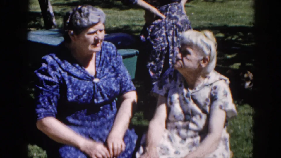 Two older women in 1960s-era floral and patterned dresses seated on a grassy lawn in Illinois, appearing to talk animatedly.