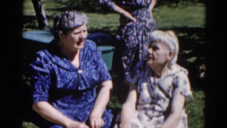 Two older women in early-1960s dress seated on a grassy lawn in Illinois, in close conversation, with characteristic 8mm film grain visible.