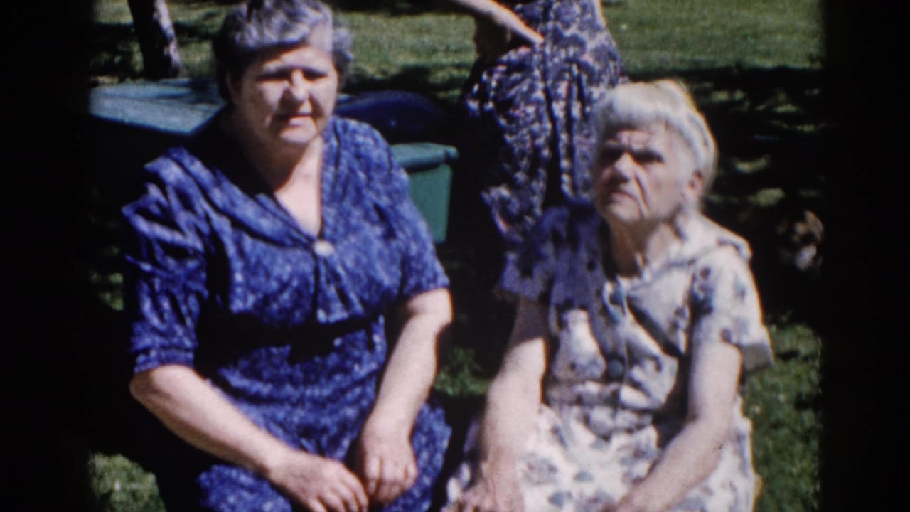 Two older women in early-1960s patterned house dresses seated in a suburban backyard, a third woman partially standing behind them, Illinois circa 1962.