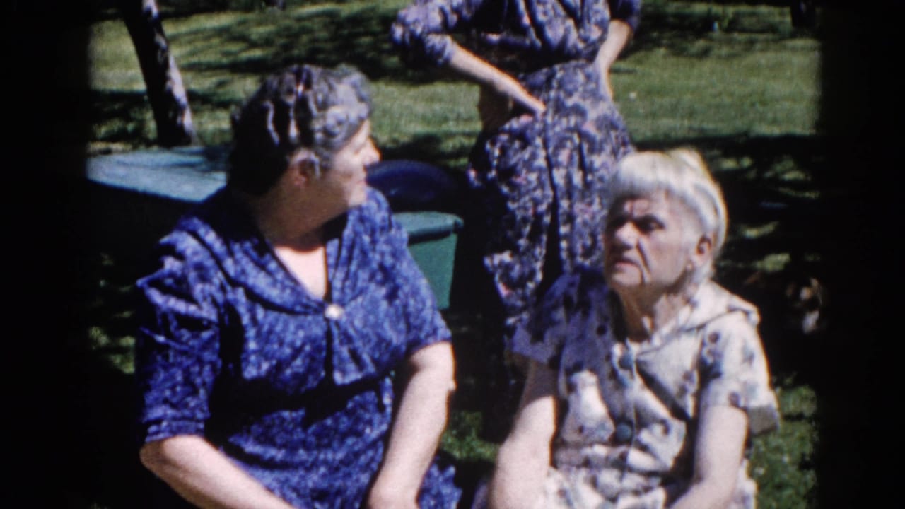 Two women in patterned dresses near dark green outdoor furniture, a third figure partially visible at upper right, suburban outdoor setting.
