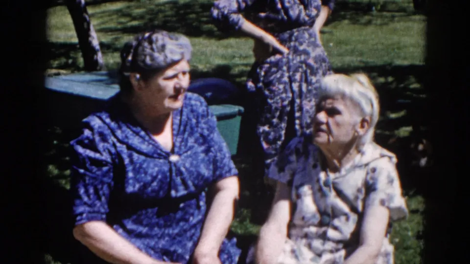 Two women in 1960s house dresses — one blue patterned, one lighter floral — in candid conversation in a suburban Illinois backyard, circa 1962.