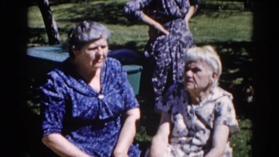 Two older women in early-1960s patterned house dresses seated in a sunlit suburban backyard, consistent with Illinois home-movie footage circa 1962.