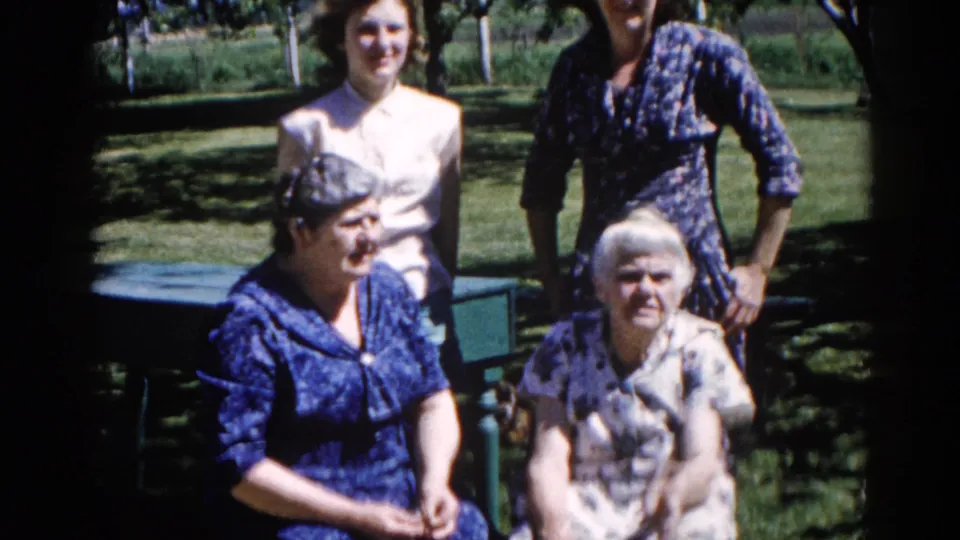 Close final frame of three women from the same Illinois family group, c. 1962; elderly woman seated on a garden bench, flanked by two younger relatives in mid-century dresses.