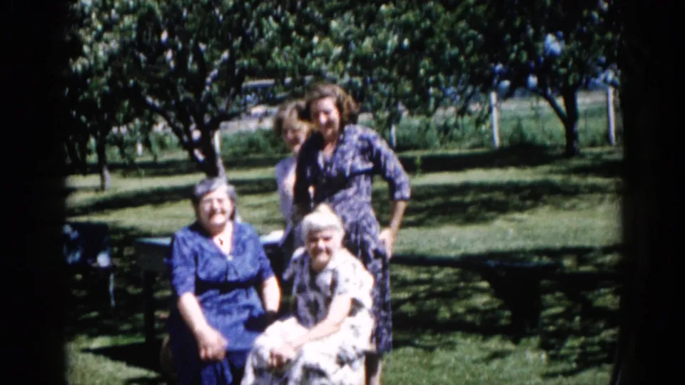 Three women in a residential garden, circa 1962 Illinois; camera appears closer as group settles into informal pose; mature trees and lawn visible.
