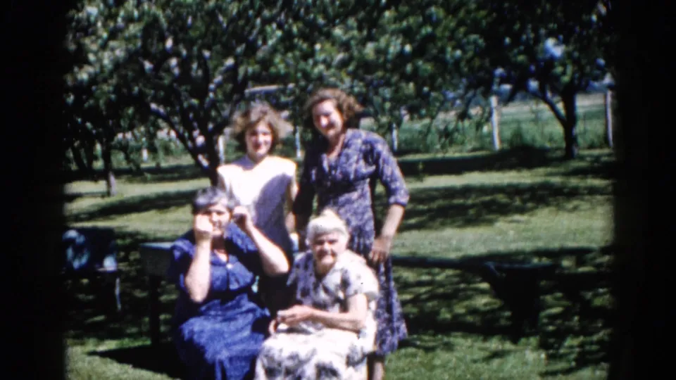 Three women posed informally in a residential garden, likely Illinois circa 1962; warm 8mm film grain and color shift visible; women engage with the camera.