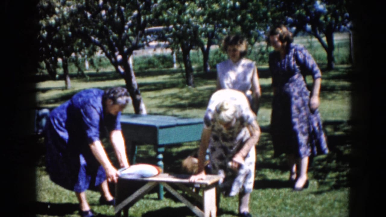 Two women lean over a green picnic table outdoors as a third woman in a blue dress stands to the right.
