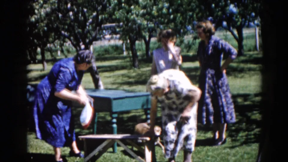 Two women in early-1960s dress stoop near a green park table in a sunlit outdoor setting, Illinois circa 1962.