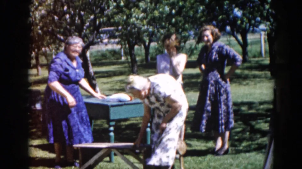 An elderly woman in a 1960s patterned dress is assisted toward a seat at a green park table by two companions, Illinois circa 1962.