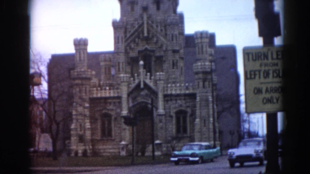 Gothic Revival stone building with crenellated towers; turquoise 1960s sedan and yellow traffic sign in foreground.
