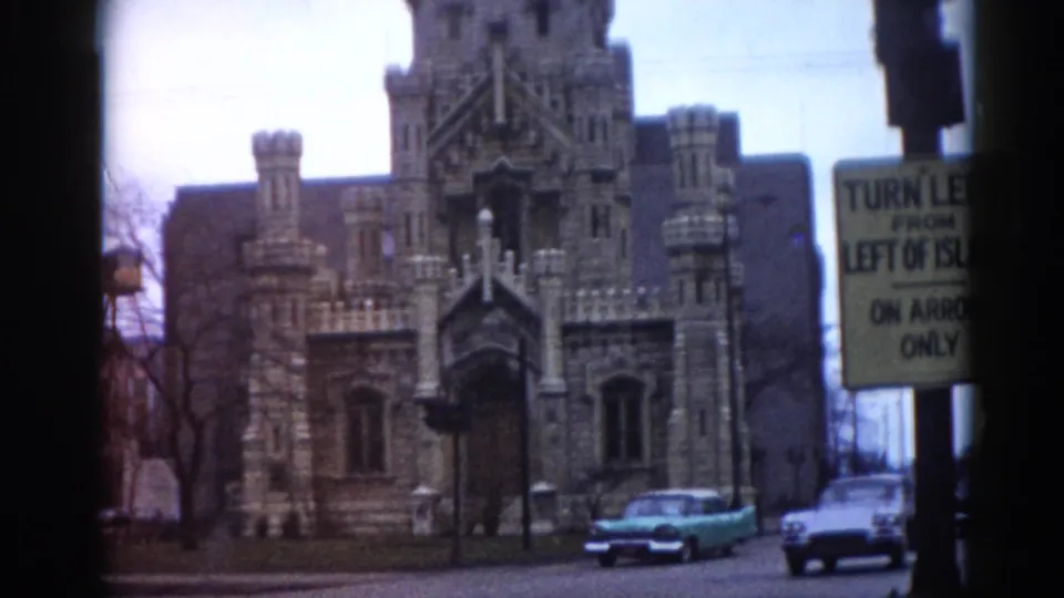 Gothic Revival institutional building with crenellated towers and ornate stonework; turquoise early-1960s American sedan and yellow traffic sign; Illinois street, 1962, 8mm home movie.