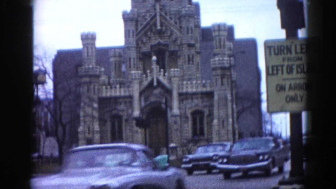 Gothic stone building in background; grey car passing in motion blur with turquoise sedan behind; traffic sign partially legible.