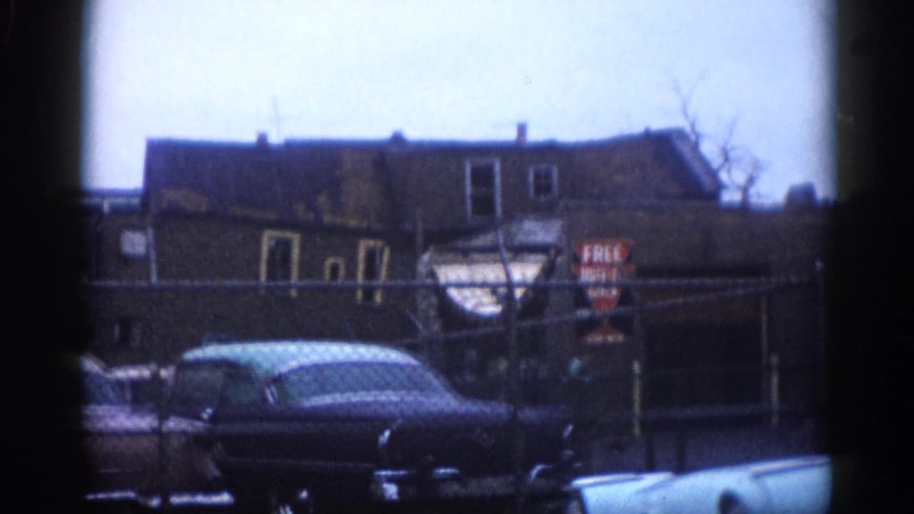 Multi-story brick building with chain-link fence, parked early-1960s American cars, and visible commercial signage in an Illinois neighborhood, circa 1962, shot on 8mm color film.