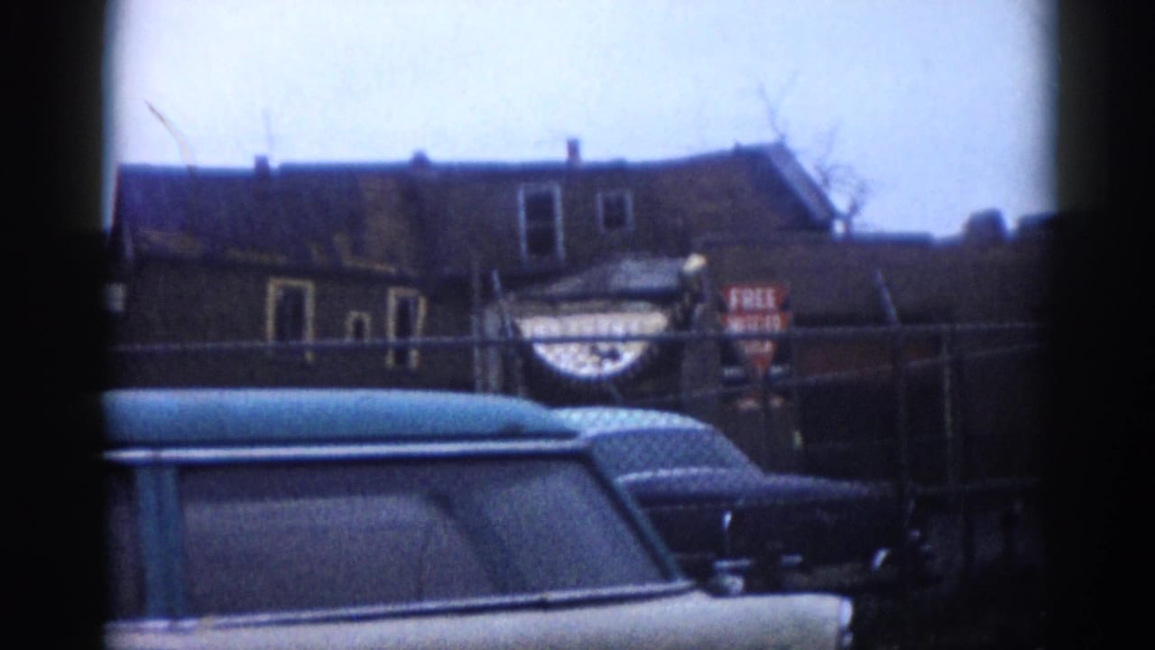 Light blue early 1960s station wagon parked near a chain-link fence, with a brick building and legible commercial signage visible behind it.