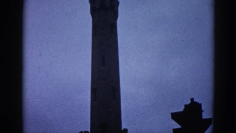 Mid-section of a tall masonry civic tower against a pale overcast sky in Illinois, circa 1962; a small sculptural or architectural element is visible at lower right, 8mm color film.