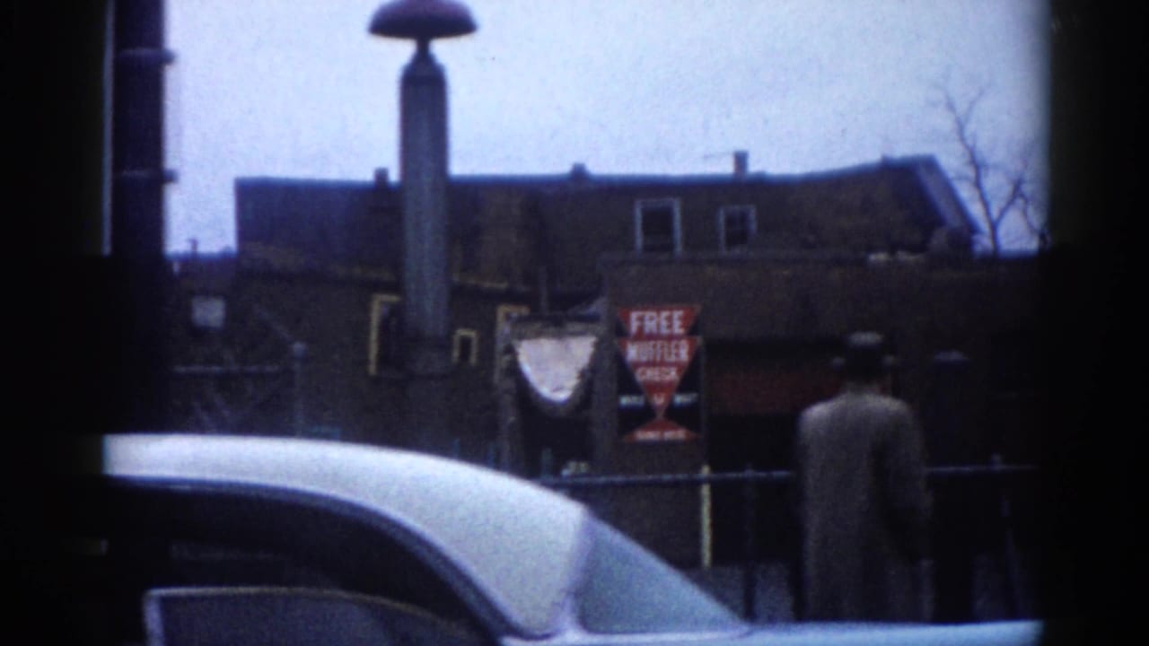 Close view of a light car roof near a street pole; FREE MUFFLER CHECK sign on a brick building and a pedestrian in dark coat visible behind.