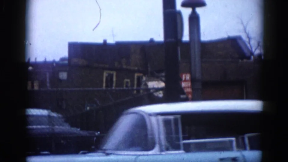 View from beside a moving vehicle on an Illinois commercial strip circa 1962: large sedan roof, brick buildings, chain-link fencing, and bare winter trees.