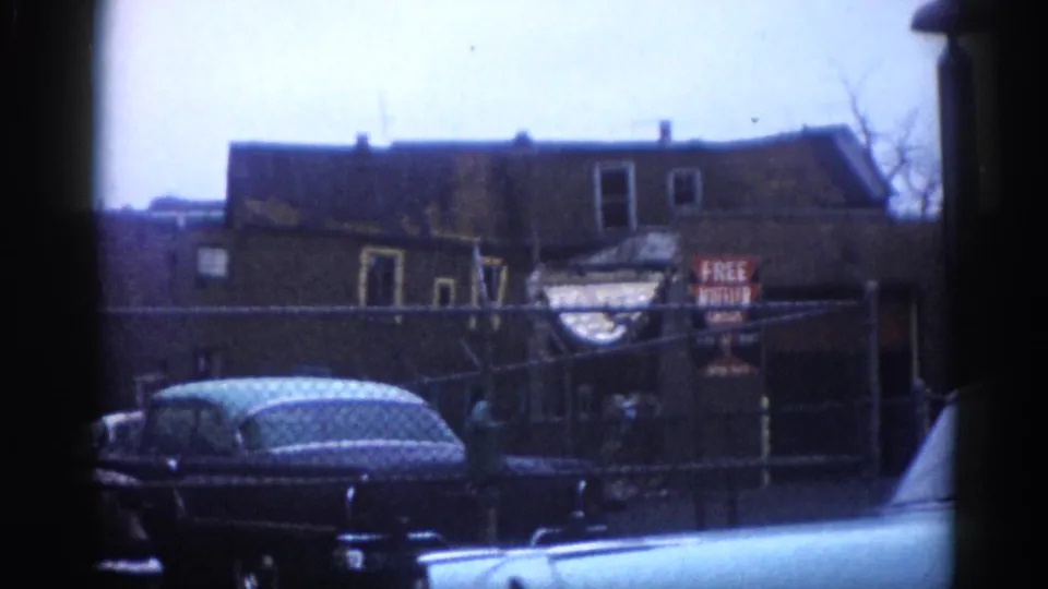 Moving street-level view on an Illinois commercial strip circa 1962: dark American sedan passing brick storefronts with a recurring FREE MUFFLER CHECK sign.