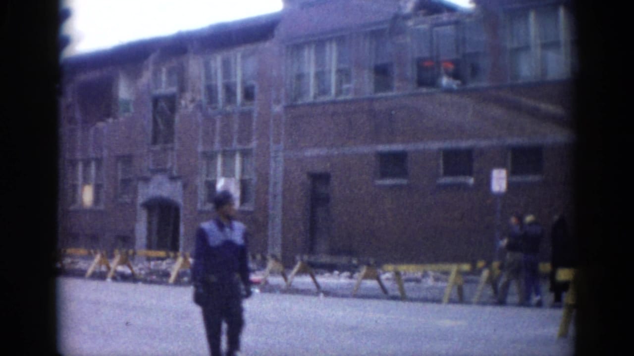 Uniformed man walks along a street in front of a multi-story brick building with broken windows.