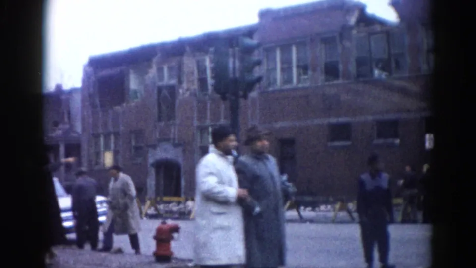 Civilian bystanders in winter overcoats gathered on a sidewalk before multi-story damaged brick buildings, urban scene consistent with Illinois circa 1962.