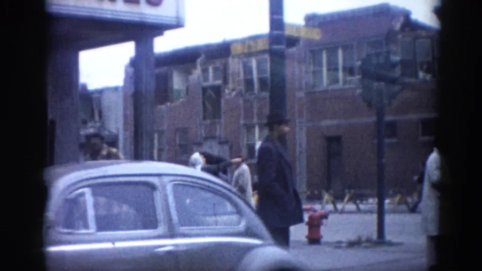 A vintage rounded sedan on a city street alongside a bystander in coat and hat; multi-story damaged brick buildings with broken windows visible behind, circa early 1960s Illinois.